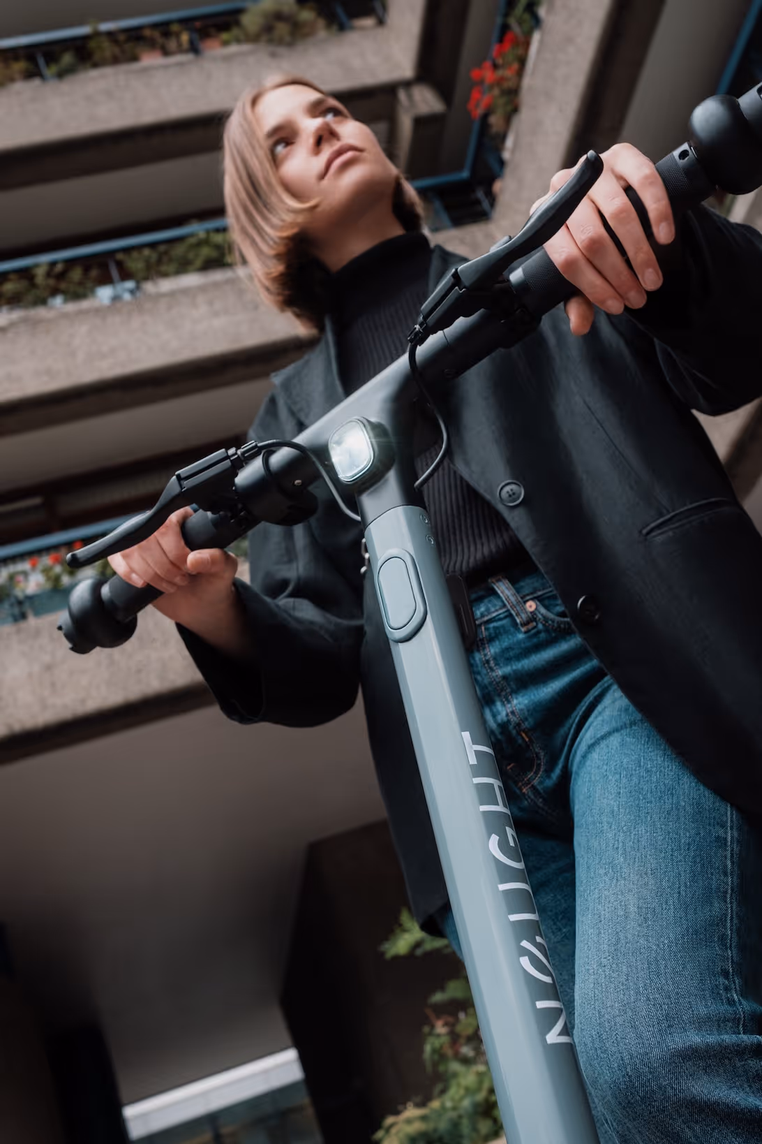 Young woman wearing a black jacket and jeans riding a gray electric scooter from a low angle.